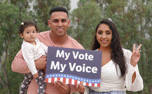 A Young Hispanic Family Smiles For The Camera While Holding Up A Sign Saying "My Vote My Voice".  