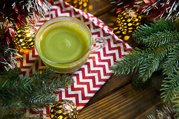 Green Japanese matcha tea in a transparent mug in Christmas decorations on a napkin with a zigzag pattern. Wooden table, spruce branches, garland, new year, comfort, warmth. Space for text