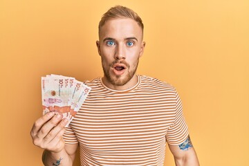 Young caucasian man holding 10 colombian pesos banknotes scared and amazed with open mouth for surprise, disbelief face