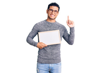 Wall Mural - Hispanic handsome young man holding blank empty banner surprised with an idea or question pointing finger with happy face, number one