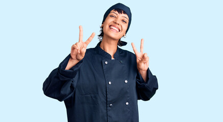 Young brunette woman with short hair wearing professional cook uniform smiling looking to the camera showing fingers doing victory sign. number two.