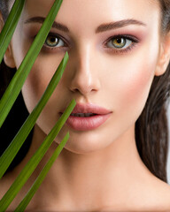 Wall Mural - Young beautiful woman with healthy skin of face and palm leaves. Closeup fresh face of an attractive caucasian girl with green plants. Model with bright brown eye makeup. Skin care concept.