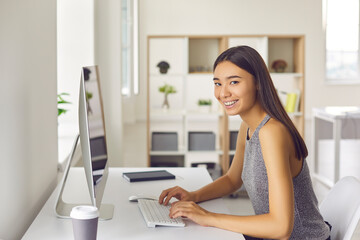 Young woman smiling and looking at camera while sitting at desk and working on computer