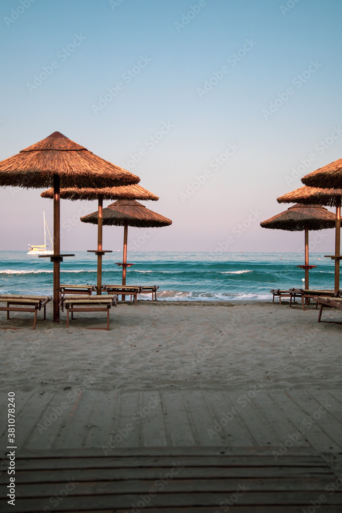 Beautiful dusk and blue hour light just after sunset in a beach club in Villasimius, Sardinia, italy. Straw sub umbrellas and wooden sunbeds, soft pink sky, summer magic atmoshpere. - obrazy, fototapety, plakaty 