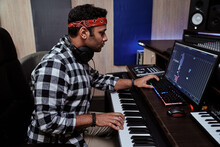 Young Man, Male Artist Looking Focused While Playing Keyboard Synthesizer, Sitting In Recording Studio