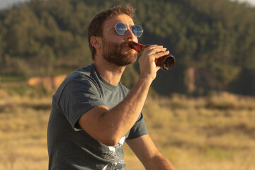a young man, with glasses and a beard, drinks beer from a bottle, while enjoying the landscape and t