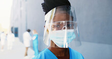 Portrait African American Beautiful Woman Doctor In Medical Mask, Face Shield And Goggles Looking At Camera. Close Up Female Physician In Respiratory Protection. Doctors On Background. Protected Suit.