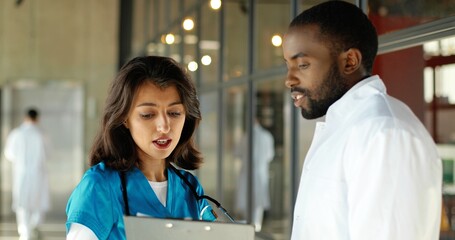 Mixed-races male and female doctors talking joyfully in clinic. Multi ethnic man and woman, medics having discussion in hospital. Physician with nurse. Doc and assistant. Conversation.