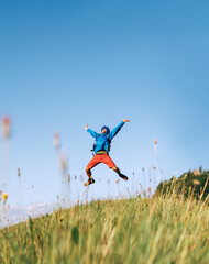 Wall Mural - backpacker traveler emotionally jumping over green grass mountain meadow with backpack with wide ope