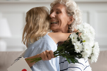 Lighting from happiness. Laughing mature grandmother or elderly foster mom folding to breast small kid girl grandchild or adopted daughter receiving greeting card and flowers on Mothers Day or March 8