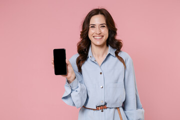 Wall Mural - Smiling young brunette woman 20s wearing casual blue shirt dress posing hold mobile cell phone with blank empty screen mock up copy space isolated on pastel pink colour background, studio portrait.