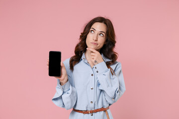 Wall Mural - Pensive young brunette woman 20s in casual blue shirt dress hold mobile phone with blank empty screen mock up copy space put hand prop up on chin isolated on pink colour background, studio portrait.