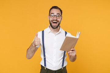 Wall Mural - Excited young bearded man 20s wearing white shirt suspender eyeglasses posing standing holding in hands reading book showing thumb up isolated on bright yellow color wall background, studio portrait.