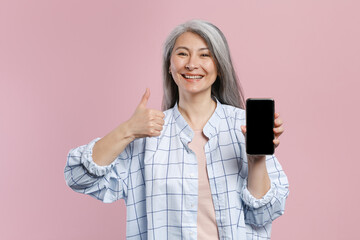 Canvas Print - Smiling gray-haired asian woman in white checkered shirt showing thumb up hold mobile cell phone with blank empty screen mock up copy space isolated on pastel pink colour background, studio portrait.