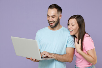 Smiling cheerful young couple two friends man woman 20s wearing blue pink empty blank design t-shirts standing working on laptop pc computer isolated on pastel violet color background studio portrait.