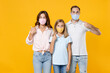 © ViDi Studio - Young parents mom dad with child kid daughter teen girl in basic t-shirts pointing index fingers on face mask to safe from coronavirus virus covid-19 isolated on yellow background. Family day concept.