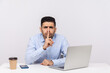 © khosrork - Shh, be quiet! Businessman sitting office workplace with laptop on desk, asking for silence or secrecy, keeping finger on lips and hushing, confidential information. indoor studio shot isolated