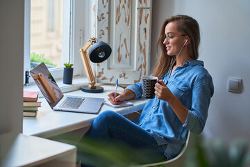 Casual young smiling smart woman student in headphones satisfied with learning foreign language. Female making notes at notebook during watching webinar video courses. Online education