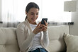 © fizkes - Happy millennial girl sit relax on couch at home using modern smartphone gadget, browsing internet. Smiling young Caucasian woman rest in living room text or message on cellphone, shopping online.