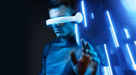 Young man in futuristic costume on dark background. Guy using VR helmet. Augmented reality, virtual reality, future technology, game concept. Blue neon light. 