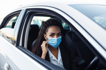 Young business woman behind the steering wheel of a taxi adjusts the medical mask. Business trips during pandemic, new normal and coronavirus travel safety concept.