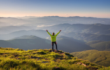 Sporty man standing on the mountain peak with raised up arms against mountain valley in fog at sunset in autumn. Happy young man, rocks, forest and blue sky in fall. Traveler hiking in mountains