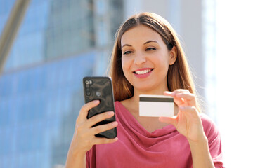 Wall Mural - Close up of delighted nice woman holding credit card and using smartphone outdoors