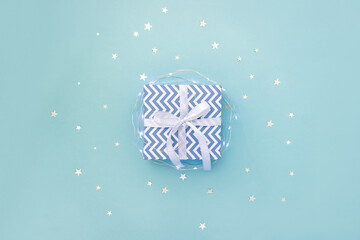 Present box with blue chevron pattern and white bow on pastel background surrounded by light garland and star confetti. Minimal concept of Christmas. Top view, flat lay