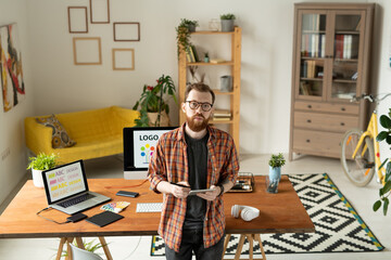 Wall Mural - Serious creative designer standing by table with computer monitor on background