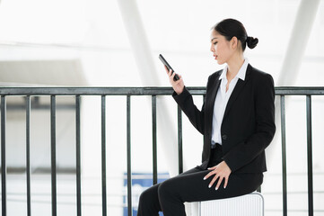 Wall Mural - Young asian business woman sitting on trolley and using smartphone. Beautiful female in black suited.