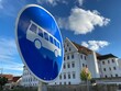 Blaues Verkehrszeichen, Bus abgebildet, dahinter historisches Gebäude mit vielen Fenstern, blauer Himmel, weiße Wolken