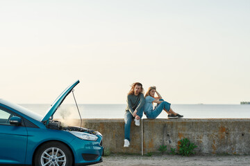 Wall Mural - Outdoor shot of couple of travelers sitting near the broken car with open hood and smoking engine, man and woman looking tired, frustrated while waiting for assistance