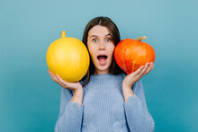 Portrait Of Emotional Attractive Young Woman With Open Mouth, Holding Two Small Pumpkins Near Face, Surprised Looking At Camera, Dressed In Sweater, Isolated On Blue Studio Background. Autumn Concept