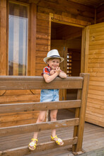 Cute Little Girl In A Cowboy Hat Hanging On A Fence Of A Wooden House.