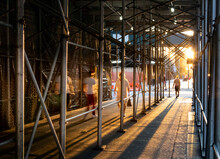 People Walking Down A Sidewalk In New York City With The Light Of Sunset Shining Through Construction Scaffolding Overhead