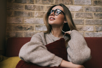 Wall Mural - Contemplative hipster girl with pillow in hands resting at comfortable sofa while thinking,pensive female student in optical eyeglasses for provide vision correction feeling pondering in cafe interior