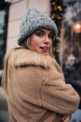 Canvas Print - Half length portrait of youthful female tourist 20 years old in trendy apparel looking at camera during winter photo session, charming hipster girl dressed in stylish coat and knitted winter hat