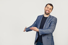 Young Businessman Man With A Beard In A Jacket, Inviting To Enter, Smiling Naturally With An Open Hand. Portrait Of A Man On A Gray Background