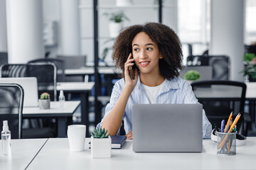 Wall Mural - Working with modern technology and clients remotely. Friendly african american lady talking on phone with client, sitting at table with laptop in office interior