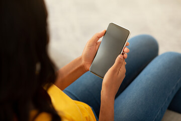 Wall Mural - African American lady holding smartphone with empty screen whileresting on sofa at home, mockup for mobile app design