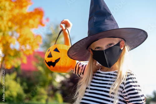 Halloween kids mask. Portrait blonde girl in witch costume with pumpkin bucket. Child wearing black face masks outdoors protecting from COVID-19.