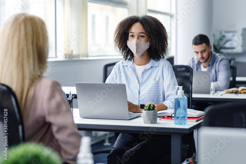 Work with client after quarantine and social distancing. Friendly african american woman in protective mask talking to european lady, sitting at workplace with laptop