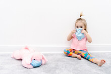 Little Blond Caucasian Girl Baby In Facial Protective Mask Sits At Home At Quarantine With Pink Dog Toy In Facial Protective Mask, To Protect From Spread Of Contagious Corona Virus Infection,