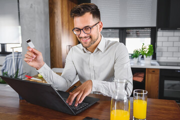 Young man holding credit card sitting in front of laptop computer at home, paying for online order. People, lifestyle, modern technologies and e-commerce concept. Online banking and shopping using mod