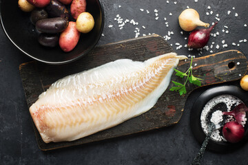 Cod white fish potatoes dish ingredients for healthy comfortable home food. Raw white fish fillet in a baking dish on a dark background. Top view.,