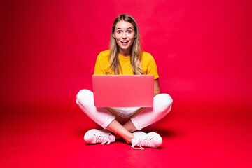 Happy young excited woman sitting on the floor with crossed legs and using laptop on red background.