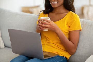 Wall Mural - Unrecognizable black lady watching entertainment video on laptop computer while drinking coffee on sofa at home