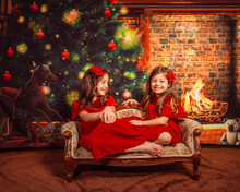 Christmas Portrait Of Two Little Smiling Girls Wearing Red Costumes On Christmas Tree Background Sitting On A Vintage Bench