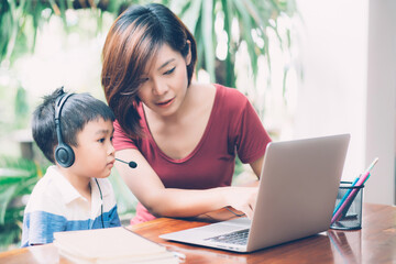 Wall Mural - Young asian mother and son using laptop computer for study and learning together at home, boy wearing headphone for e-learning with distancing, teacher or mom support child, education concept.