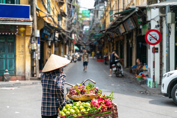 The street vendor with bike loaded of tropical fruits in old town street in Hanoi, old houses and street activites on background